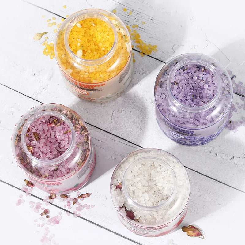 Quatre pots en verre contenant des sels de bain colorés dans les tons jaune, violet, rose et blanc.